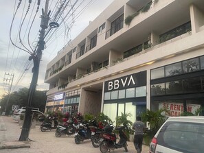 A multi-story building with a sign for 'BBVA' on the ground floor. There are several parked motorcycles and a few cars. - Modern 2BR Apartment in Tulum Great Location (Tulum)
