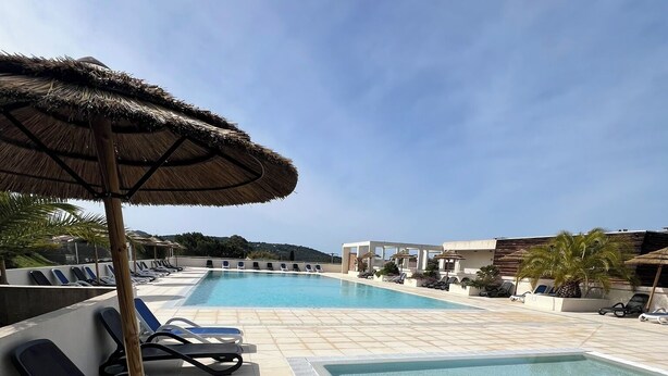 Un área junto a la piscina con sillones de descanso, una sombrilla de paja y una piscina azul cristalina.