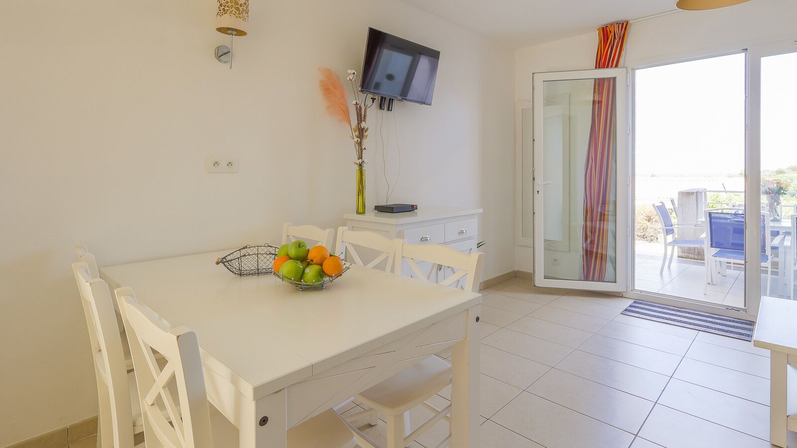 Un coin repas avec une table blanche, des chaises, un saladier de fruits, un téléviseur et une porte-fenêtre coulissante donnant sur un espace extérieur aménagé avec des chaises.