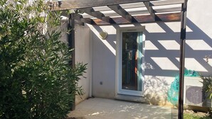 Un área de patio techada con una pérgola de madera, una puerta que da a una habitación y un pequeño jardín con plantas verdes.