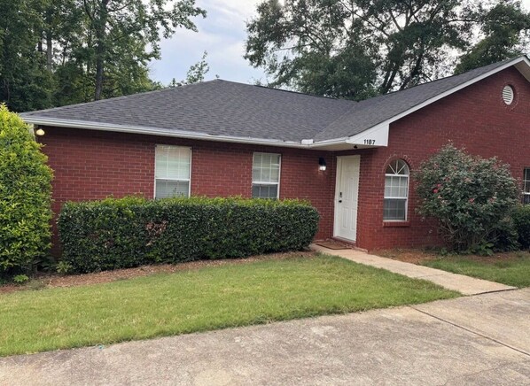 A single-story brick house with a white door and windows, a small front yard with a hedge, and a number '1187' on the house. - 3 Free Parking  Home in Auburn Al (Auburn)