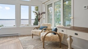 A room with a large window overlooking a body of water, two upholstered chairs with cushions, a wooden desk, and a woven rug. - Oceanfront Acadia Views Arcade Game Room Pets Ok (Surry)