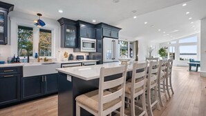 A modern kitchen with dark cabinetry, a central island with bar stools, and a large window providing a view of the outdoors. - Oceanfront Acadia Views Arcade Game Room Pets Ok (Surry)