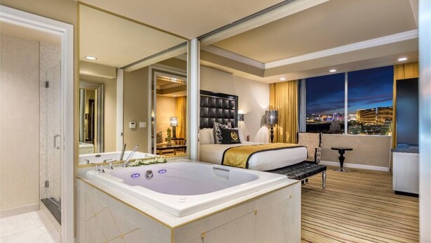 A modern hotel room with a large bathtub, a bed with a gold accent, a city view, and a balcony with a table and chair.
