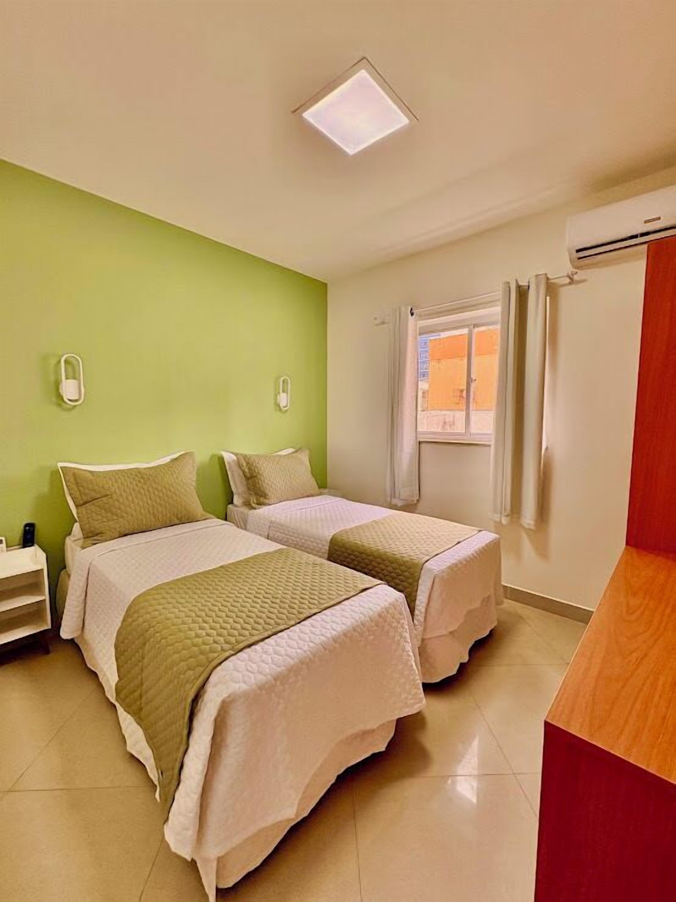 A hotel room with two beds, a small desk, a window with curtains, and a ceiling light.