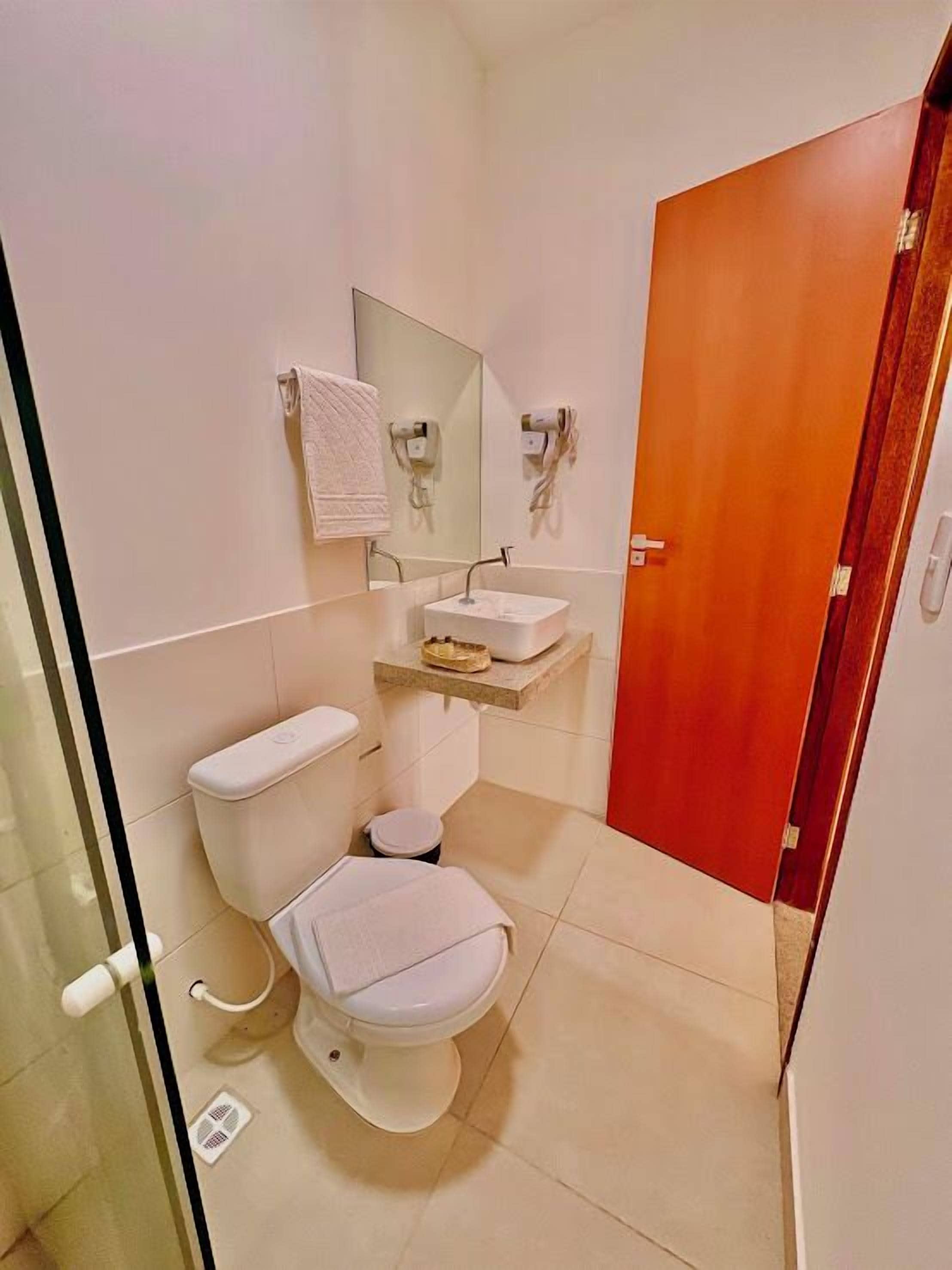 Een kleine badkamer met toilet, wastafel en spiegel erboven. Een handdoek hangt aan de muur en rechts is een deur.