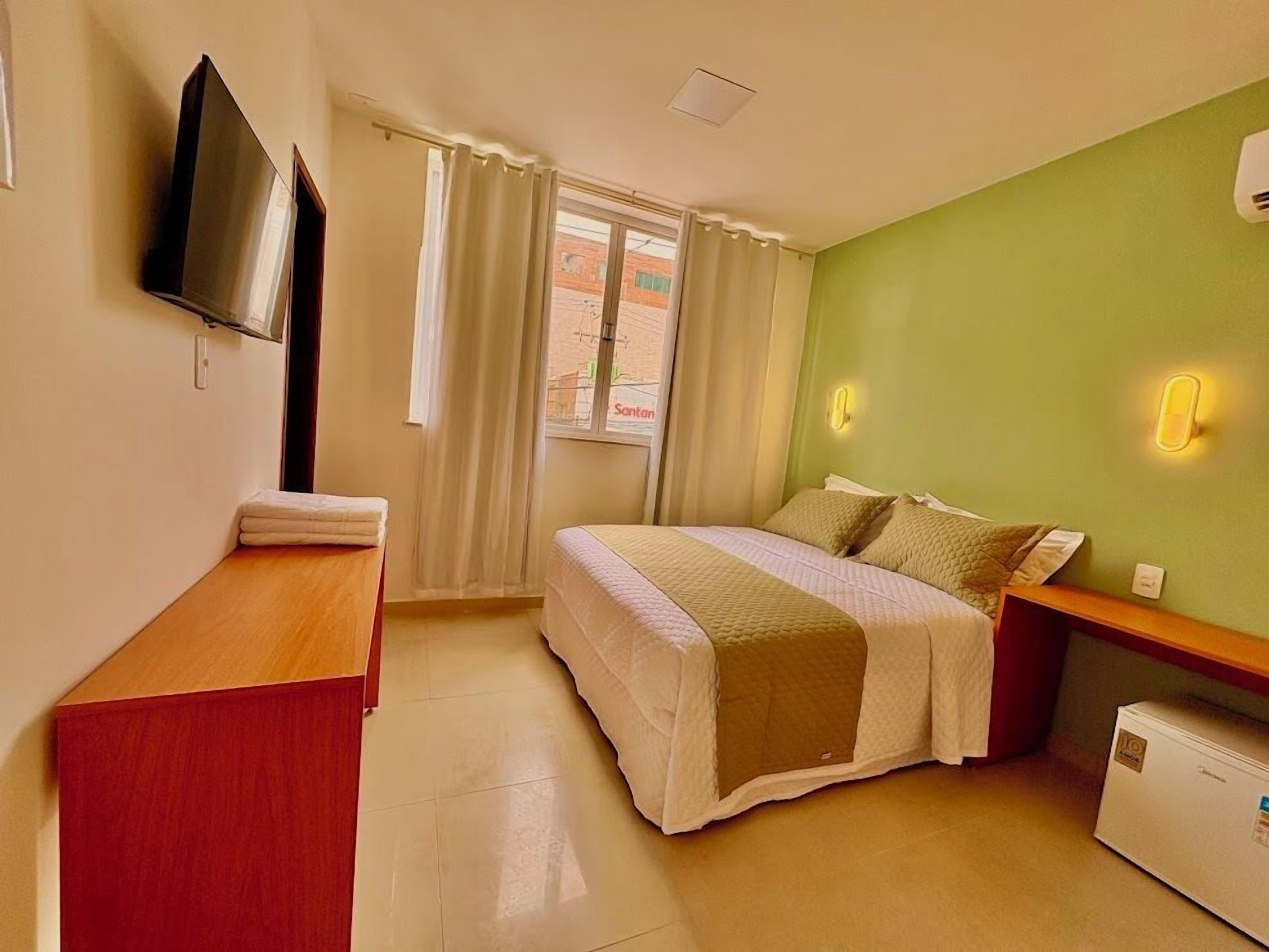 A hotel room with a bed, a desk, a TV, a mini-fridge, and a window with curtains.