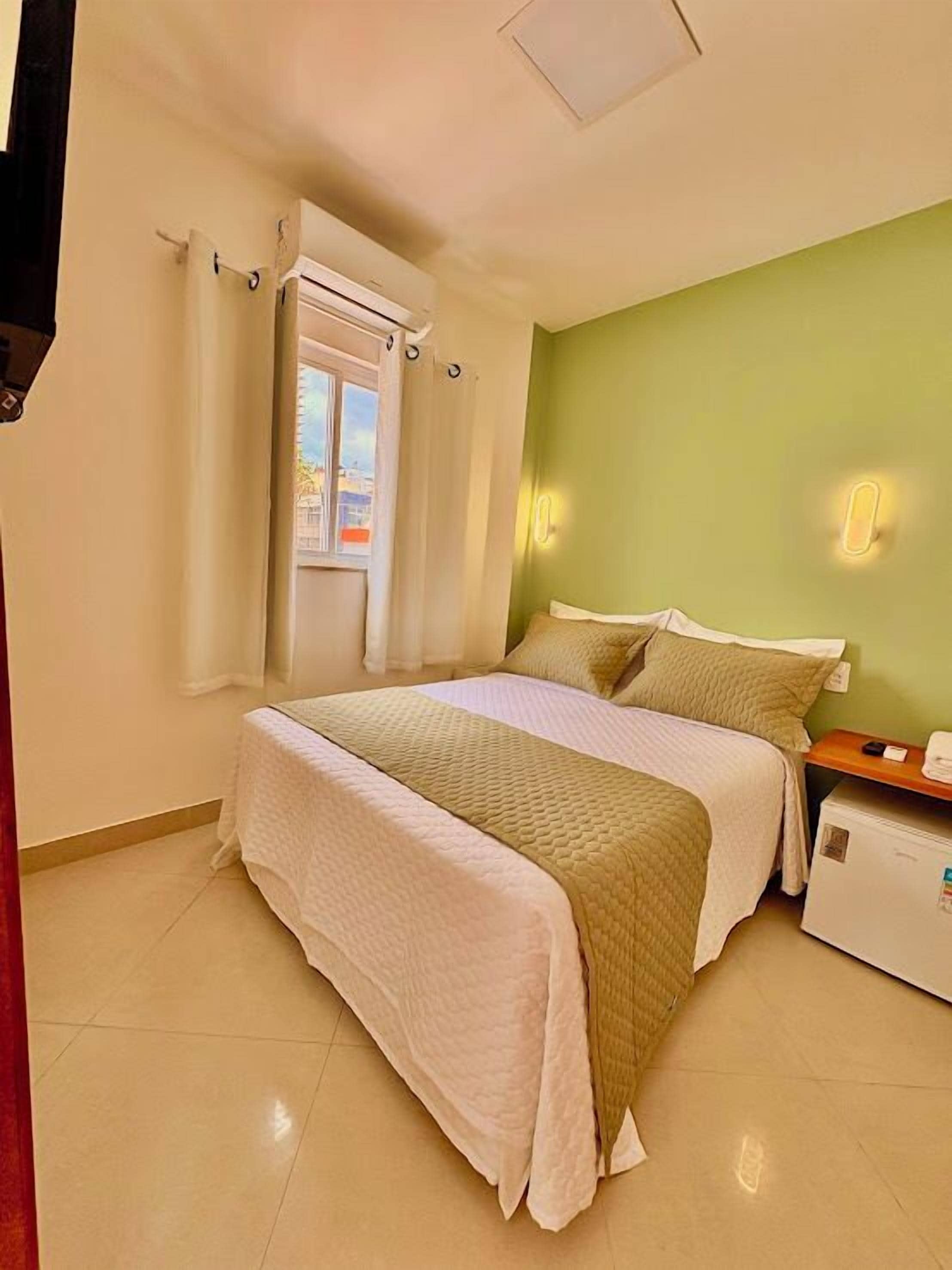 Un dormitorio con dos camas, una ventana con cortinas, una lámpara de pared y un refrigerador pequeño.
