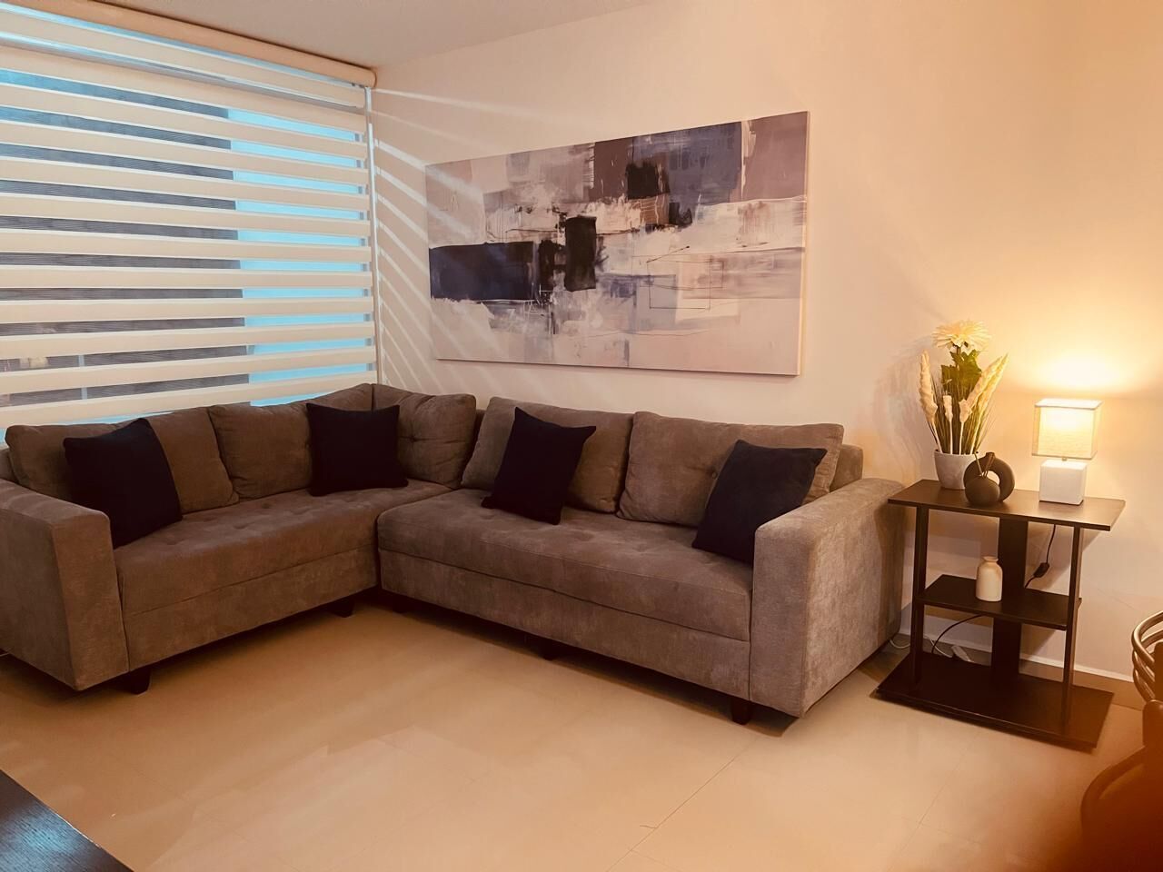 A living room with a sectional sofa, a modern abstract painting, a side table with a lamp, and a window with blinds.