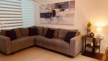 Een woonkamer met een modulaire bank, een modern abstract schilderij, een bijzettafeltje met een lamp en een raam met jaloezieën.