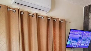 Ein Zimmer mit beige farbenen Vorhängen, einem wandmontierten Flachbildfernseher, einer kleinen Sitzbank und einem Bett mit dunkler Bettdecke.