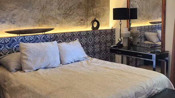 Ein Schlafzimmer mit einem Bett, einem Deckenventilator, einem Nachttisch mit Lampe und einer Wand mit strukturierter Oberfläche.