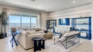 A spacious living room with a large window offering a view of the sea, a blue bookshelf, and a comfortable seating area with a sofa and chairs. - Luxe Desert Escape w/ Strip View - 360° Las Vegas Strip & Mountain Views (Henderson)