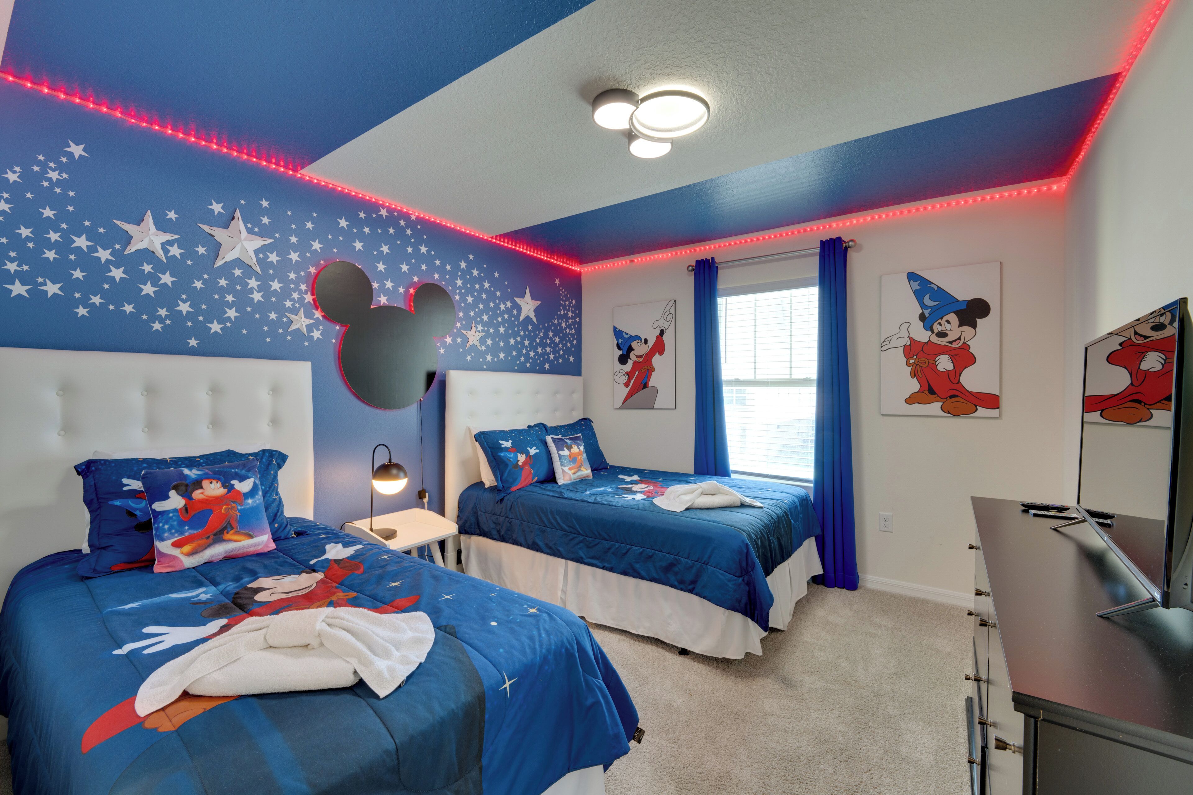 Een kamer met twee bedden, wanddecoraties met sterren, Mickey Mouse afbeeldingen en een bureau met een computer.