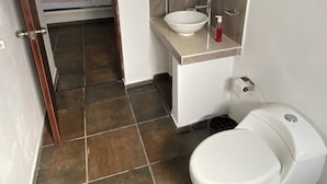 Een badkamer met een wit wastafel, een spiegel, een toilet en een bed in de aangrenzende kamer.