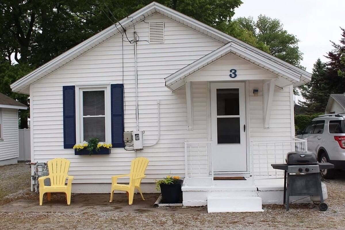 A small white house with the number 3 on the door, two yellow chairs, and a grill outside.