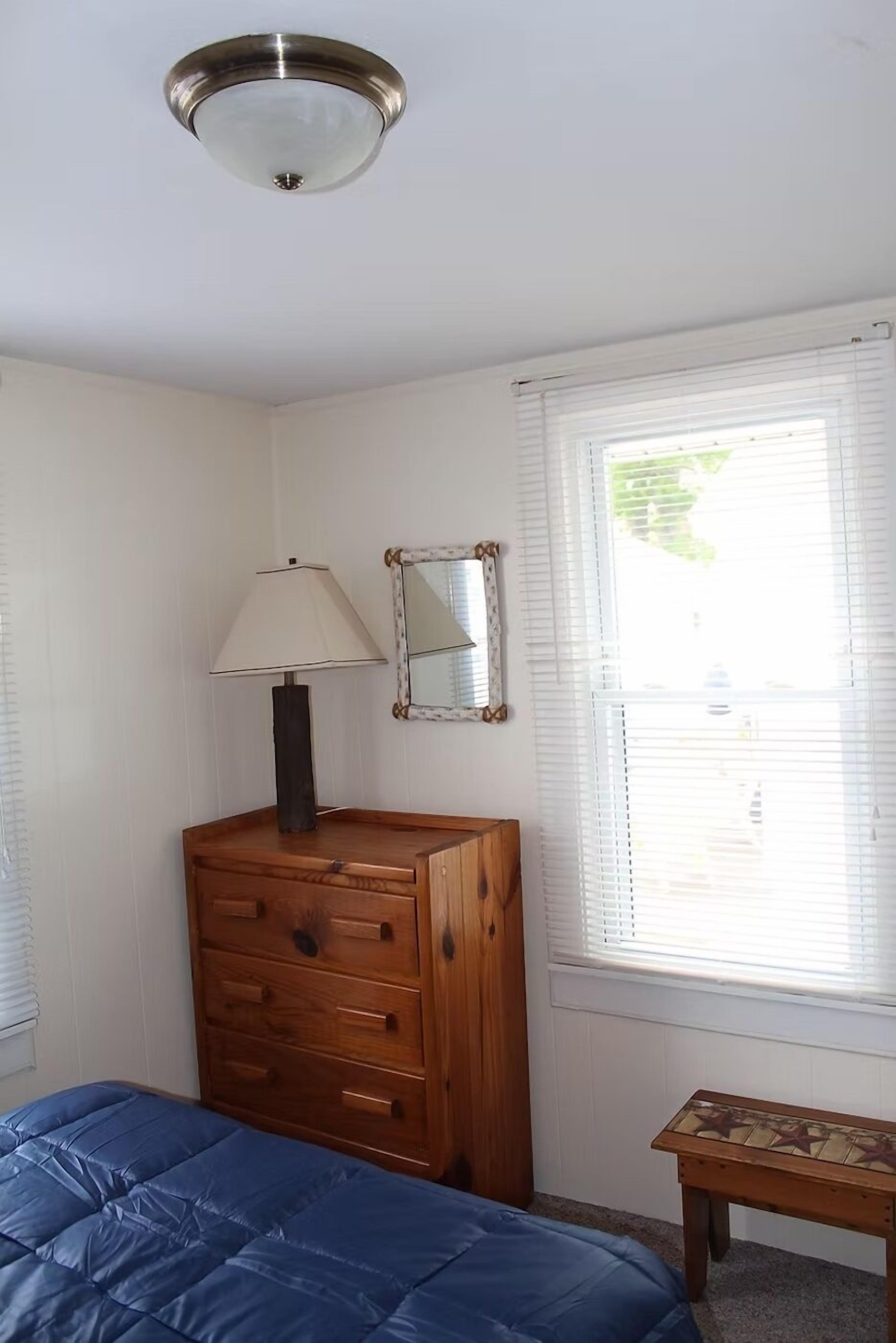 A bedroom with a bed, a wooden dresser, a lamp, a mirror, and a window with blinds.