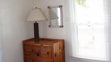 A bedroom with a bed, a wooden dresser, a lamp, a mirror, and a window with blinds.
