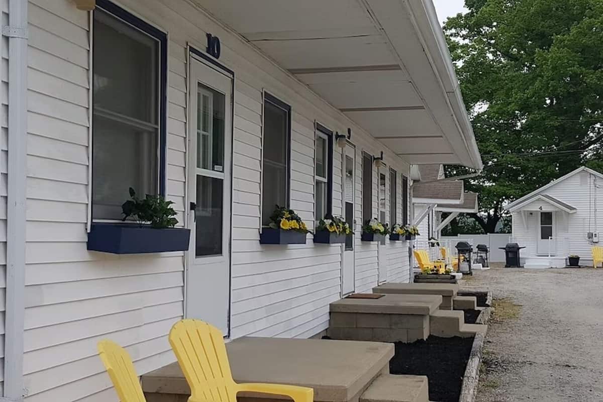 A row of motel rooms with individual windows, outdoor seating, and a small building in the background.