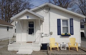 A small white house with a number '2' on the front, a porch with steps, and two yellow chairs outside. - Dockside Lake Resort (Monticello)
