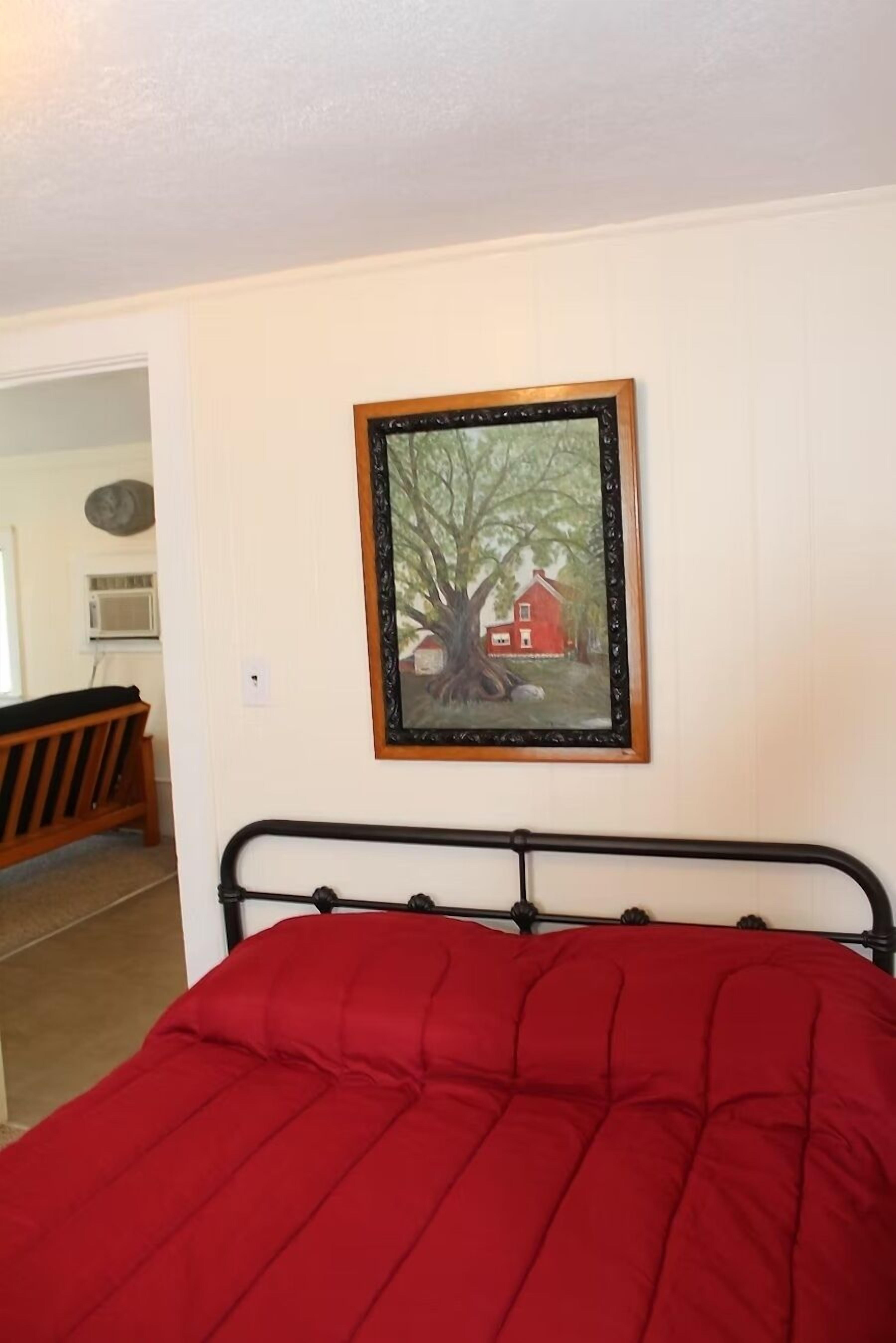 Une pièce comprenant un lit, un tableau encadré représentant un arbre et une maison, et un banc en bois.