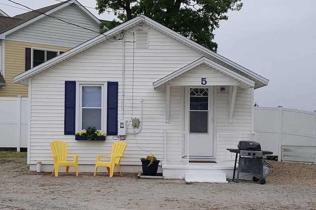 A single-story house with a white exterior, numbered 5, featuring two yellow chairs outside, a small porch, and a grill.
