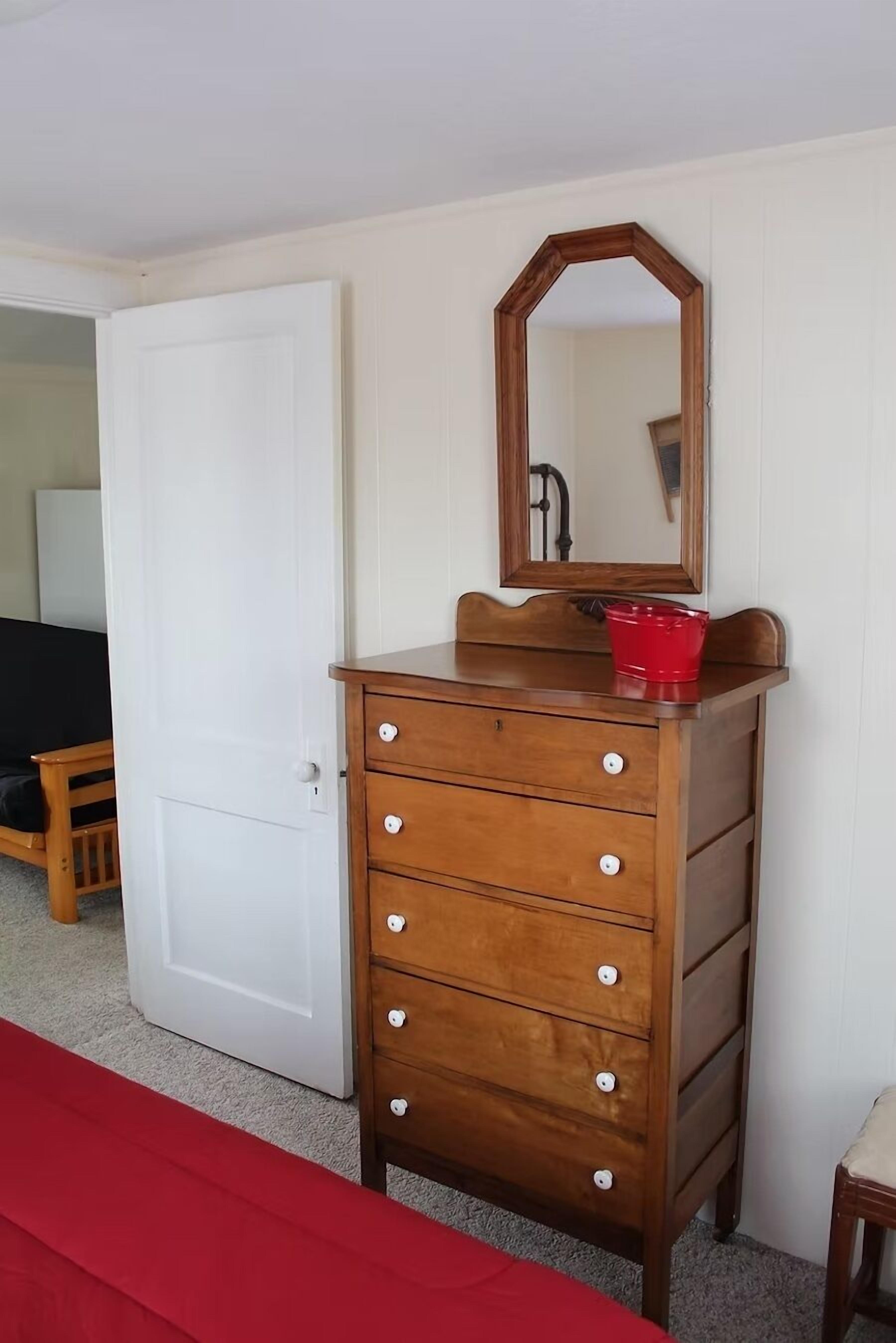 A wooden dresser with a mirror and a red bucket, a white door, and a red bench in a room with white walls.