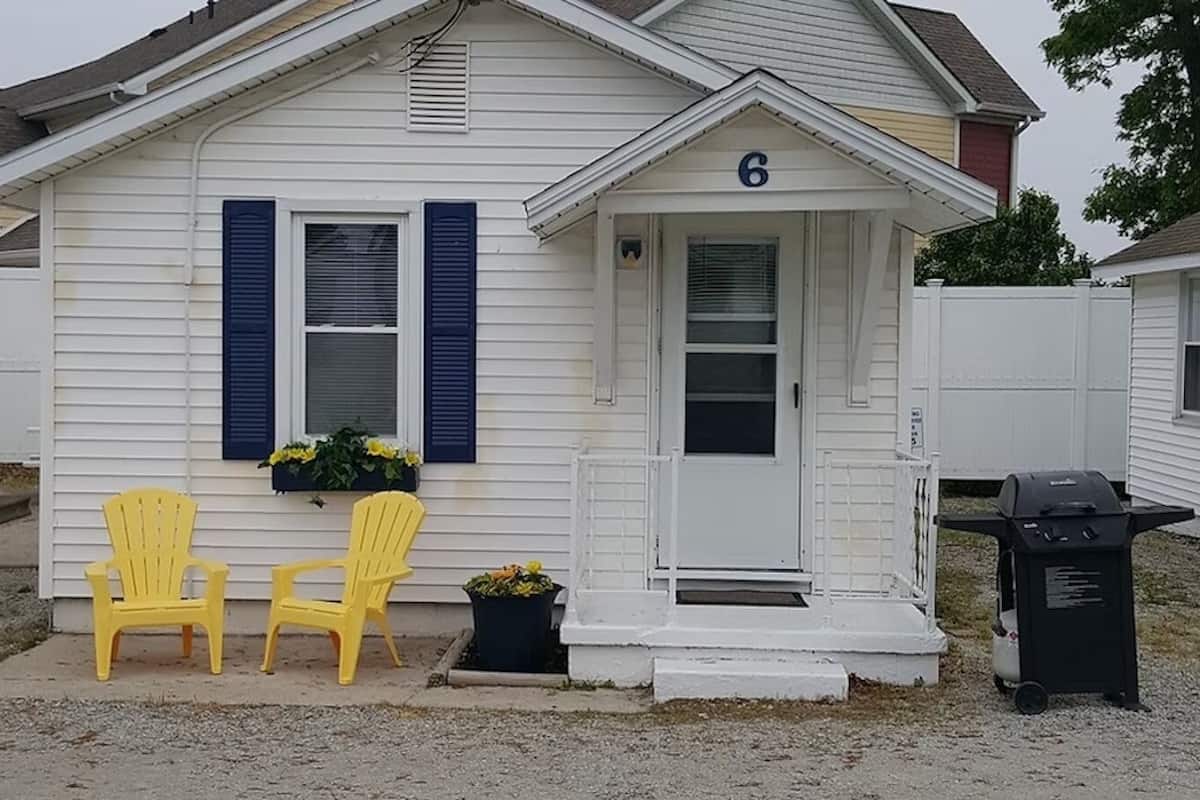 A single-story house with a white exterior and blue shutters, marked with the number 6. There are two yellow chairs outside, a small planter with flowers, and a barbecue grill on the right side.