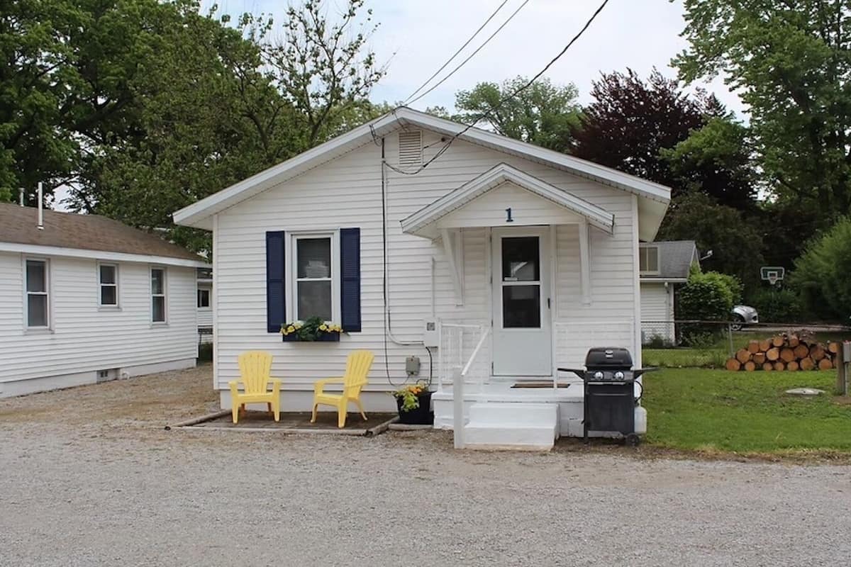 A small, single-story white house with a front porch, two yellow Adirondack chairs, and a barbecue grill.