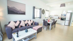Una sala moderna con un sofá gris, una mesa de centro blanca y una zona de comedor con una mesa y sillas blancas. Se observa una cocina con gabinetes blancos y un refrigerador de acero inoxidable al fondo.