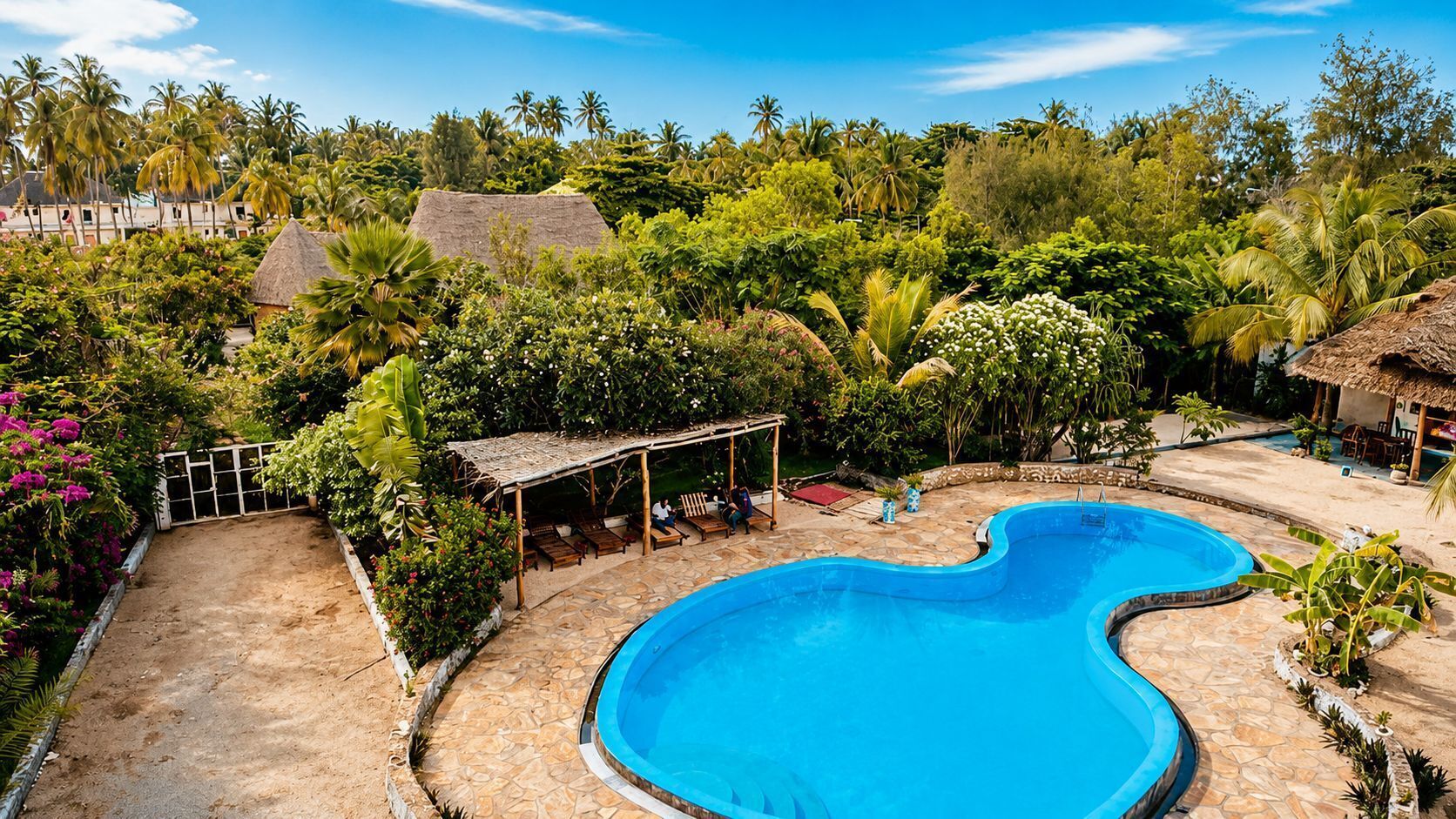 A swimming pool surrounded by lush greenery, a covered seating area, and a thatched-roof structure.