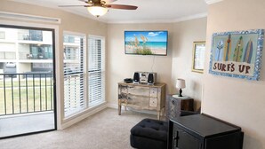 Ein Zimmer mit Deckenventilator, einem an der Wand befestigten Fernseher, einem Schreibtisch mit Lautsprechern und einem Schild mit der Aufschrift 'SURF'S UP'.