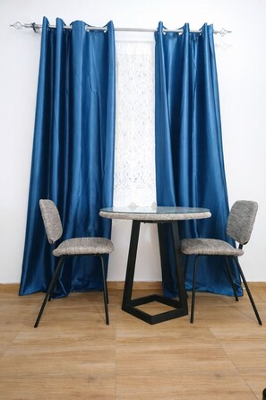 A room with a round table and two chairs, a window with a lace curtain, and a blue curtain covering the window. - CJ Apartment  (Kasoa)
