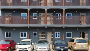 A multi-story building with balconies, a parking area with several cars, and a simple architectural design. - CJ Apartment  (Kasoa)