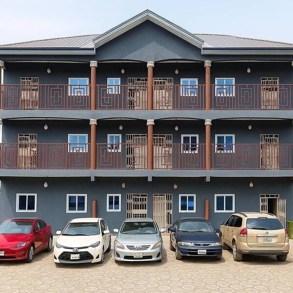 A multi-story building with balconies, a parking area with several cars, and a simple architectural design. - CJ Apartment  (Kasoa)