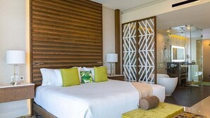 Une chambre d’hôtel moderne dotée d’un grand lit, d’une table de chevet, d’une salle de bain fermée par des parois de verre et d’un coin salon avec une petite table.