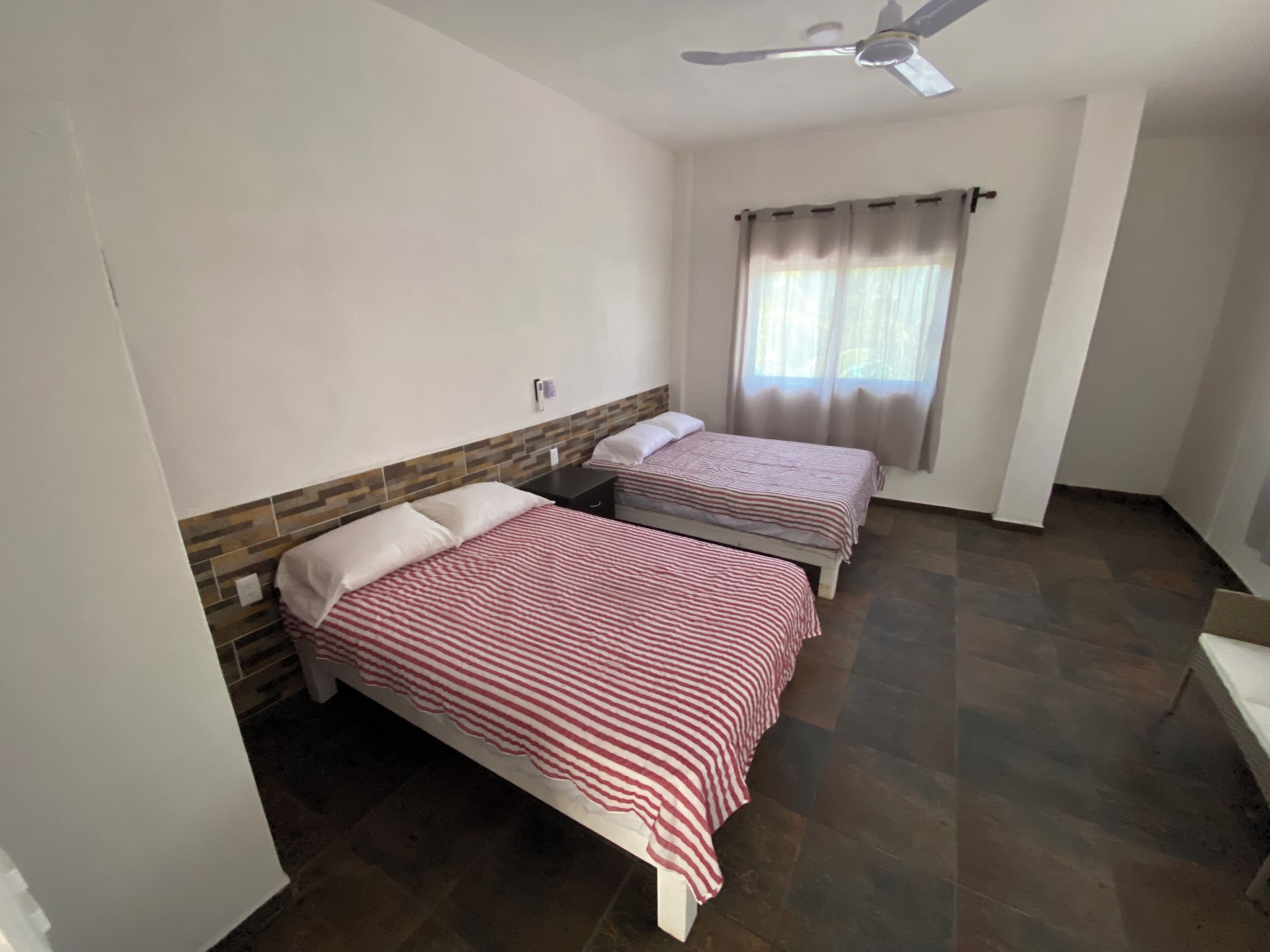 Ein Zimmer mit zwei Einzelbetten, einem Deckenventilator, einem Fenster mit Vorhängen und einem Fliesenboden.