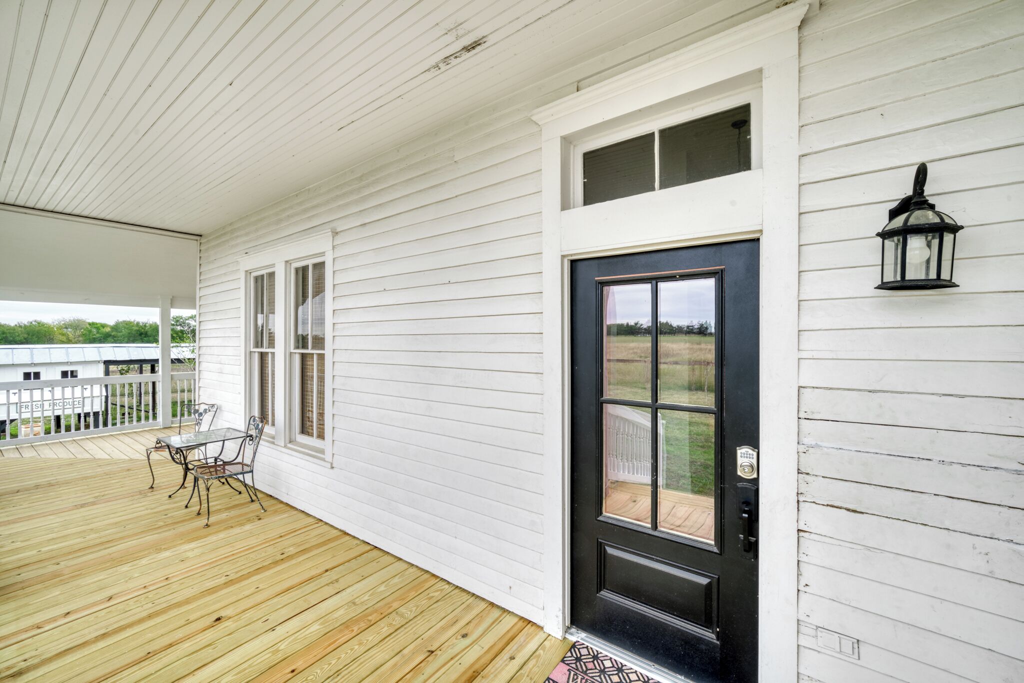 A white wooden porch with a black door and windows, a small table and chairs, and a lantern on the wall.