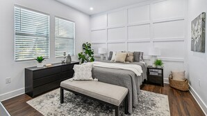 Ein Schlafzimmer mit Bett, Nachttischen, einer Bank, einem Schminktisch und einem großen Fenster mit Jalousien.