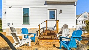 Ein zweistöckiges Haus mit einer Veranda, einem Feuerkorb aus Stein und mehreren Adirondack-Stühlen.