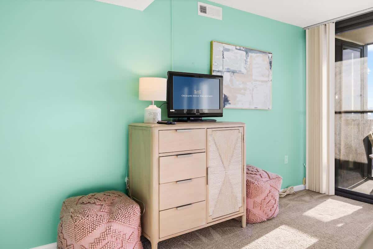 Una habitación con paredes verde azulado, un televisor sobre un mueble, un cuadro abstracto enmarcado y dos otomanas rosadas.