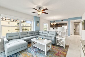 A modern living room with a sectional sofa, a white coffee table, a ceiling fan, and a view of the outside.