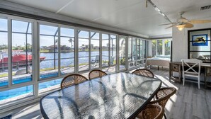 A sunroom with a large glass wall, a dining table, wicker chairs, a ceiling fan, and a view of a body of water. - Aqc15826 (Port Charlotte)