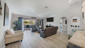 Un vaste espace de vie comprenant un canapé, des fauteuils, une table basse et un téléviseur. Un balcon aménagé avec des sièges d’extérieur offre une vue sur les palmiers.