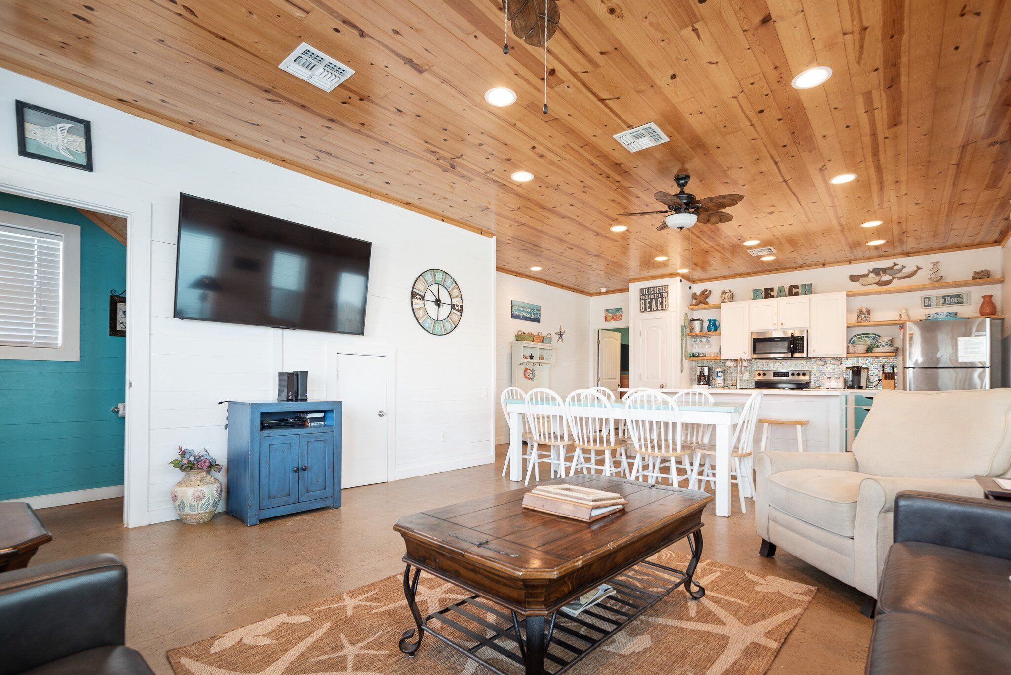 A spacious living room with a wooden ceiling, a dining area with white chairs, a kitchen with white cabinets, and a large flat-screen TV mounted on the wall.