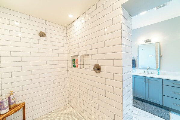 Ein modernes Badezimmer mit weißen, in Ziegeloptik angeordneten Fliesen, einer Dusche mit Handbrause, einem Waschtisch mit Spiegel und einem kleinen Tisch mit Pflegeprodukten.