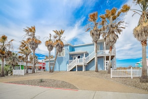 A beachfront property with a light blue building featuring a white balcony and staircase. Palm trees line the property, and a white picket fence is visible. - Villa Laguna - Pet Friendly! Beach Gear Credit Included! (Port Aransas)