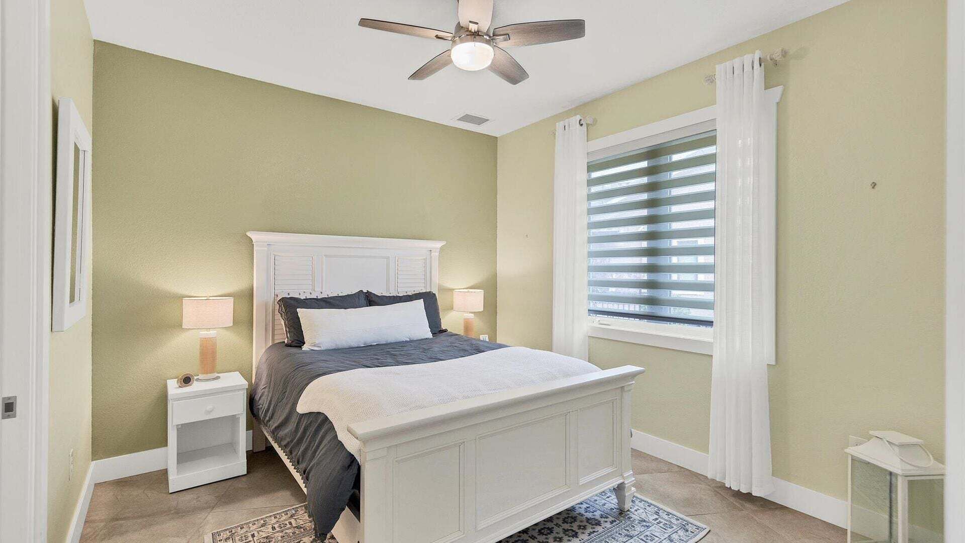 Ein Schlafzimmer mit Bett, Nachttischen, Deckenventilator, Fenster mit Jalousien und Teppich.