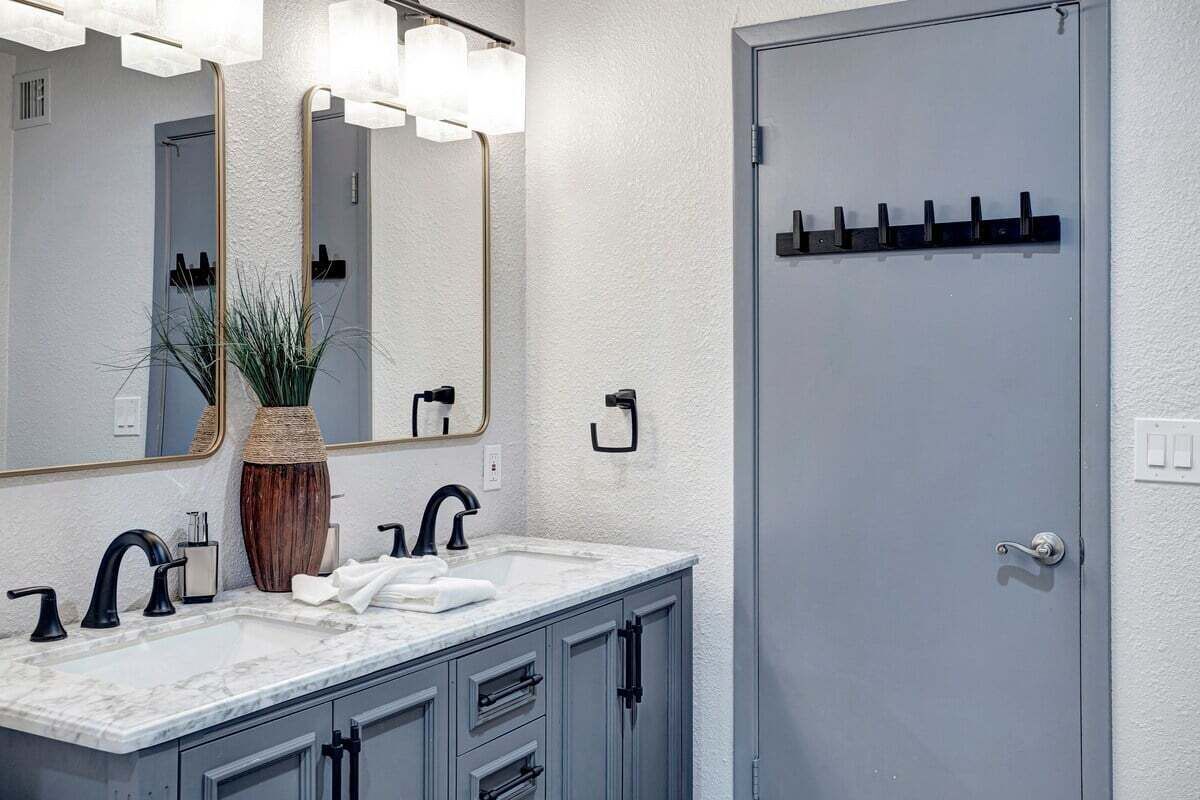A bathroom with a white marble sink, two black faucets, a large mirror with lights, a wooden vase, and a gray door with hooks.