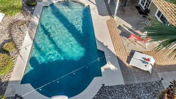 A rectangular swimming pool with clear blue water, surrounded by a paved deck. There are lounge chairs and a small table near the pool.